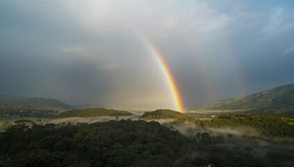 Showing primary rainbow arc rising over green hills in valley, with mist and secondary rainbow arc