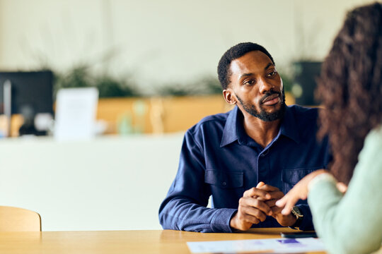 African-American Man In Navy Shirt Talks With Colleague At Office Desk