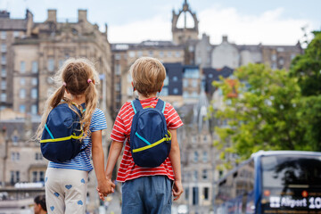 Children gaze at city traffic, holding hands with backpacks on