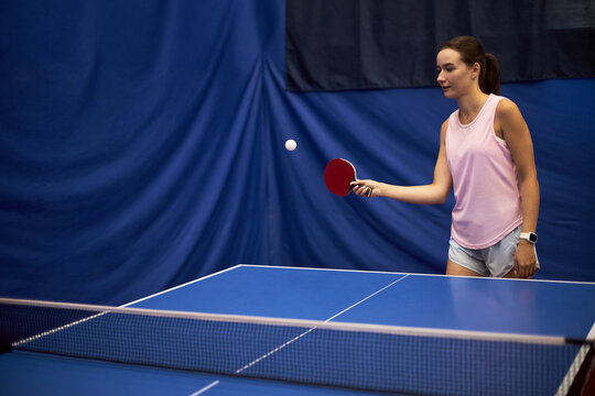 Young adult Caucasian woman playing table tennis indoors, holding paddle and focusing on ball during game, standing near blue ping pong table with blue curtain background