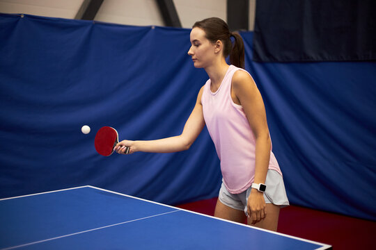 Young adult Caucasian woman playing table tennis, holding paddle in right hand and focusing on ball during game, standing next to blue ping pong table in indoor sports facility - Powered by Adobe