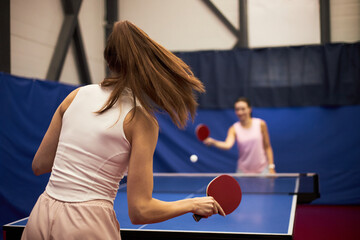 Two Caucasian young adult women playing table tennis indoors, both holding paddles and focusing on game, action captured from behind one woman with opponent visible across table
