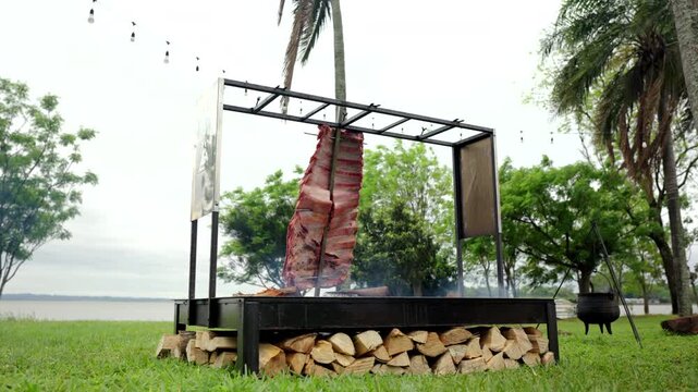Rack of costillar stands upright against glowing ember pit, framed by a custom parrilla structure surrounded by stacked firewood, smoke drifting upward in thin blue ribbons, in serene rural backdrop.