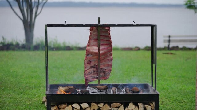 Rack of costillar stands vertically - a la cruz -, facing the heat and slowly grilling over glowing embers in a custom parrilla structure, with waters of Paran&aacute; River creating natural backdrop.