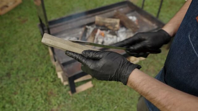 Asador pair of gloved hands holds a sharpening stone and a long knife, carefully drawing the steel against the grit, behind, the parrilla stands ready, embers glowing softly beneath stacked wood.