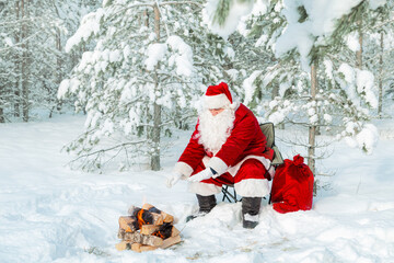 Authentic Santa Claus with a bag of gifts sits near a fire in a snowy forest.