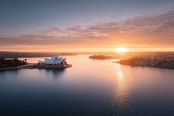 Aerial view of a city opera house at sunrise. Golden light bathes the iconic architecture, reflecting on the calm water. Perfect for travel, business, or cultural themes.