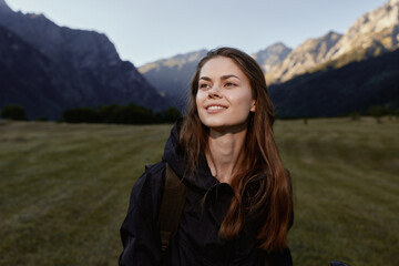Naklejka premium A cheerful young woman stands outdoors in a scenic mountain landscape. She wears a dark jacket and smiles softly, exuding calm confidence and curiosity as the setting enhances the moment.