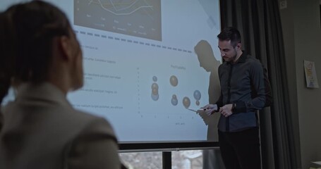 Young business associates discuss profitability projections in a meeting. The presenter points to a chart on a projector screen displaying key performance indicators. - Powered by Adobe