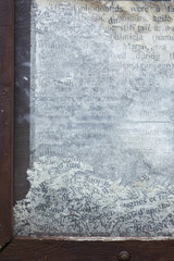 Close-up of weathered and partially erased text on a glass surface, framed by chipped dark wood. The faded black letters on a cloudy white background evoke age, decay, and forgotten information.