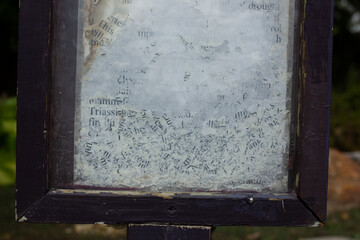 Close-up of weathered and partially erased text on a glass surface, framed by chipped dark wood. The faded black letters on a cloudy white background evoke age, decay, and forgotten information.