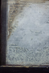 Close-up of weathered and partially erased text on a glass surface, framed by chipped dark wood. The faded black letters on a cloudy white background evoke age, decay, and forgotten information.