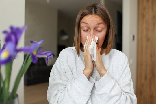 Seasonal allergy. Close up face of allergic young woman uses paper tissues from runny nose, sneezes from flowers pollen at home. Girl with close eyes with flu, itching or cough, allergy rhinitis