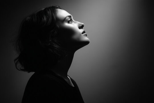 a dramatic black and white side portrait of a woman looking left and upward, strong contrast lighting from above, chiaroscuro style, minimal background, emotional and mysterious expression. horizontal
