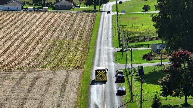 Aerial view of driving american ambulance vehicle on rural road between farmland in USA. Aerial tilt up wide shot. Flashing lights of vehicle. Sunny summer day in Virginia.
