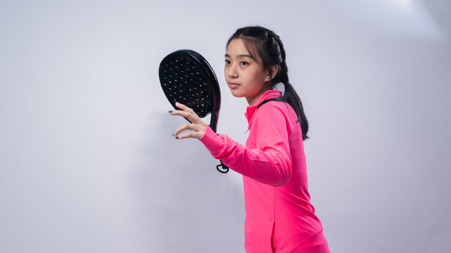 Asian woman in a pink long-sleeve sports outfit holding a black padel racket in a ready-to-play stance, looking focused and confident against a clean white studio background - Powered by Adobe