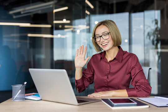 Happy businesswoman wearing glasses sitting at a desk in a modern office, having a video conference on her laptop and smiling while greeting someone with a wave