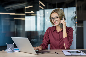 Businesswoman working in her modern office, smiling while talking on a mobile phone and typing on a laptop, representing efficient communication and multitasking in a corporate environment