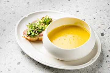 Top view of a white plate with creamy yellow soup and an open sandwich with ham and fresh greens on a light gray background. Minimalist lunch presentation, clean and modern style.