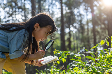 Child girl using a magnifying glass and notebook in the forest.
