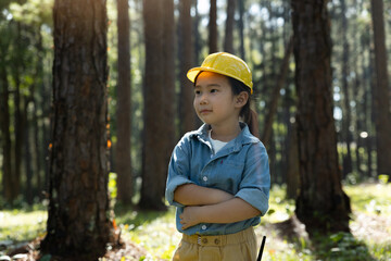 Young girl wearing yellow safety helmet standing confidently with arms crossed in forest.