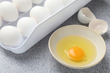 Many whole raw chicken eggs and broken one in bowl on grey table, closeup