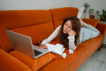 Young woman lying on a sofa in her living room, working remotely with her laptop and notebook, having a creative crisis and looking pensive, with crumpled paper balls next to her