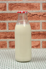 A glass bottle of milk, capped with a red and white checkered lid, stands on a green patterned tablecloth in front of a red brick background
