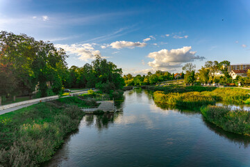 Warta River flowing through Uniejów in the Łódź Voivodeship surrounded by park greenery and urban buildings on a sunny afternoon with blue sky and clouds
