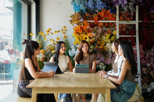 A group of women are sitting around a wooden table in a flower shop. They are all smiling and seem to be enjoying each other's company
