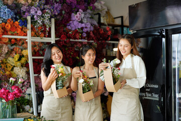 Three women are standing in a flower shop, each holding a box of flowers. The shop is filled with colorful flowers and plants, creating a cheerful and inviting atmosphere