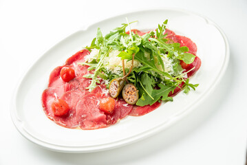 Thinly sliced raw beef carpaccio, garnished with fresh arugula, shaved Parmesan cheese, caper berries, and cherry tomatoes on a white oval plate.