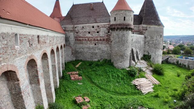 Corvin Castle, a historical landmark and Gothic-Renaissance fortress, showcasing its stone walls and tower against a backdrop of trees and town buildings under a clear sky in Transylvania, Romania
