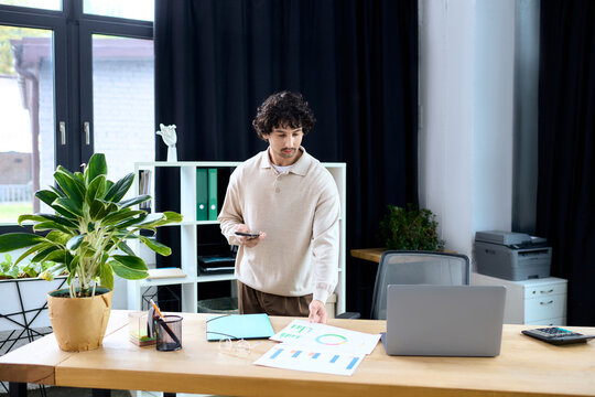 Handsome young man reviewing documents while using smartphone in modern office space