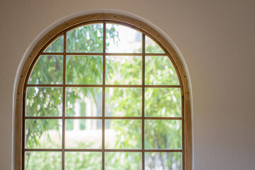 An arched window with a grid frame overlooks lush green trees.