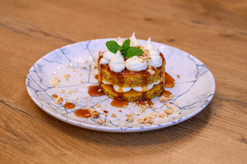 Gourmet dessert with caramel, nuts, sponge cake, and whipped cream on a marble plate, served on a rustic wooden table in fine dining restaurant setting.