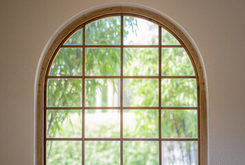 An arched window with a grid frame overlooks lush green trees.