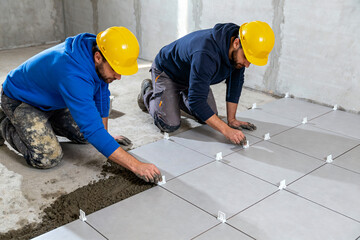 Two construction workers wearing yellow hard hats and navy blue hoodies install large gray floor tiles on a concrete subfloor, using tile spacers and cement mix during a renovation project