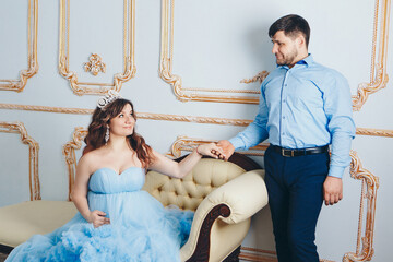 a married couple stands together near a beige sofa. couple in love looking at each other. pregnant girl in a lush blue dress. young man in a blue shirt. family photo session. 