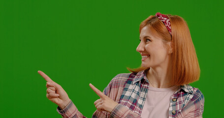 Happy woman points at commercial promotion or advertising copy space over green screen background. Portrait, product placement girl and female chroma key model on mock up background.