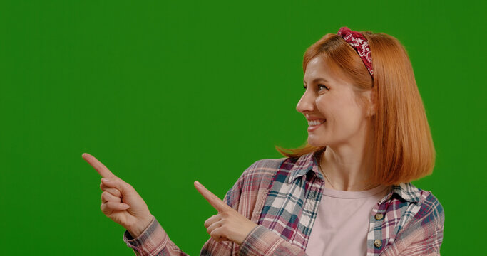 Happy woman points at commercial promotion or advertising copy space over green screen background. Portrait, product placement girl and female chroma key model on mock up background.