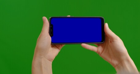 Woman hands with blue mock up chromakey screen phone closeup over green screen background. Female holds smartphone with hands watching videos.