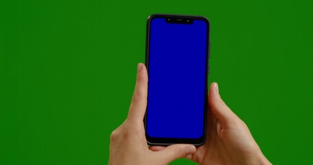 Woman holds blue chroma key screen device vertically with fingers on green background. Phone user hands watch blue chroma key screen.