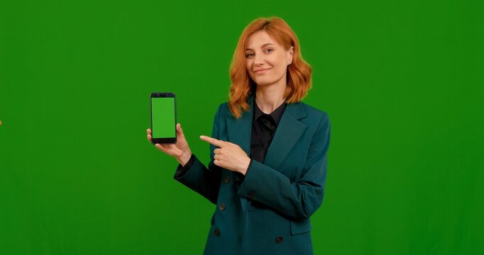 Woman pointing to mock up smartphone with aproval over green screen background. Sell advertising a product or app on chroma key phone screen.