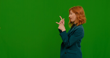 The optimistic woman points with both index fingers forward. Attractive red-haired white girl in blazer on a green screen background.