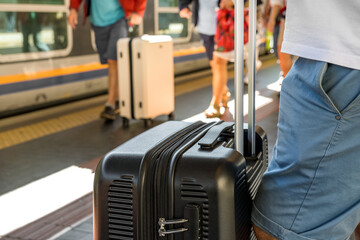 Traveler with black suitcase waiting at train station platform, surrounded by other passengers