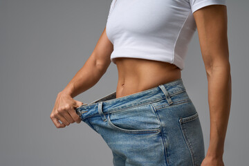 Fit woman in white crop top showing weight loss success by holding loose jeans