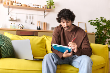 Young man writing notes while relaxing on a cozy yellow sofa in a bright living room