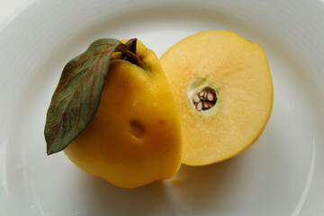 Fresh quince with green leaf and halved fruit on white plate revealing creamy interior and seeds. Bright clean food photography.