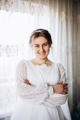 A beautiful bride in a white lace wedding dress smiles softly indoors. She stands by a window with sheer curtains, looking radiant and happy on her special day before the ceremony.